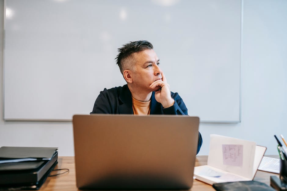 Man working on a laptop in an office looks thoughtful and tired.